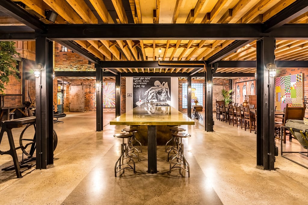 The_Compound Brass Island - Designs 05 Industrial-style interior with a large brass dining bar, featuring a polished brass countertop, metal bar stools, and exposed brick walls in a warm, ambient setting.