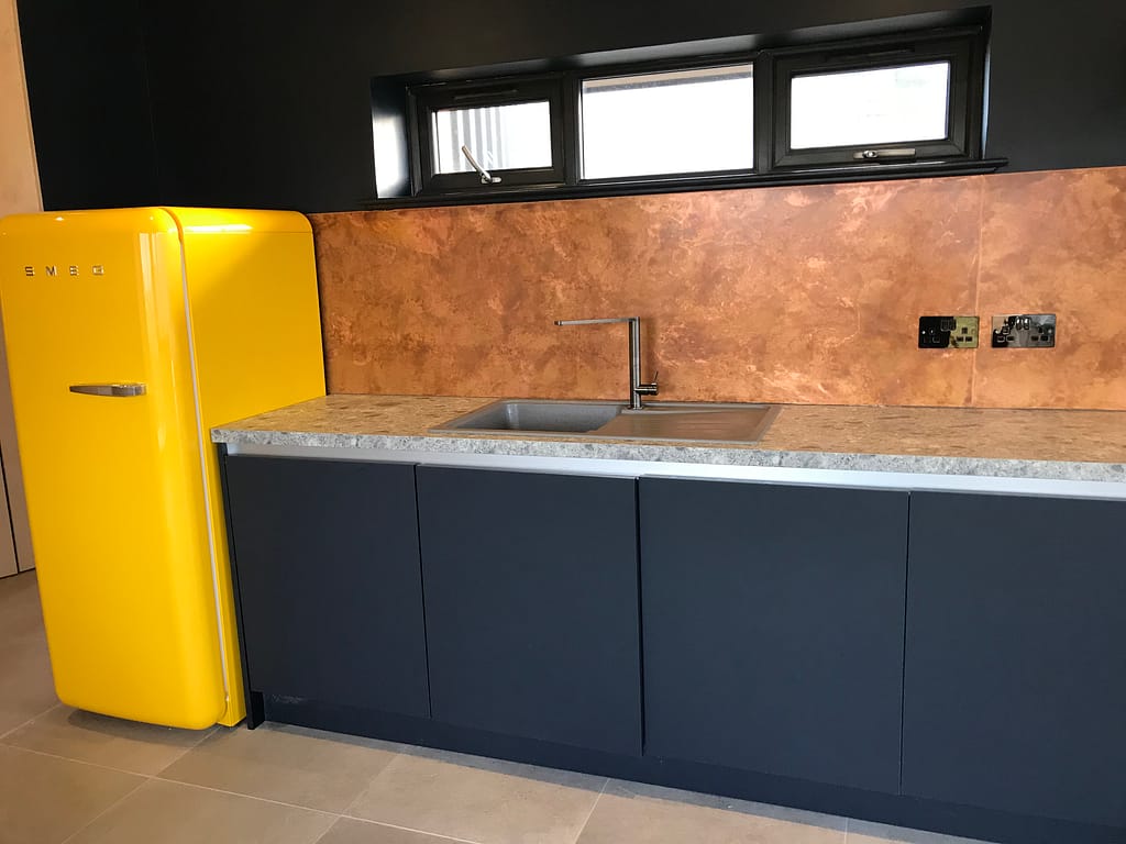 Aged Copper Kitchen Splashback with Yellow SMEG Fridge - Designs 05 Aged copper kitchen splashback with a warm patina, paired with dark cabinets, a grey worktop, and a yellow SMEG fridge.