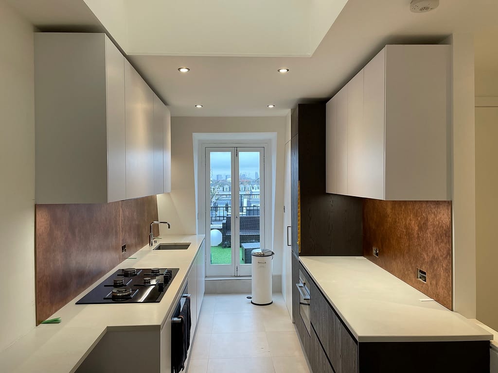 Twin Aged Copper Splashbacks in a Sleek, Modern Kitchen - Designs 05 Two matching aged copper splashbacks installed in a contemporary kitchen with white cabinetry and large glass doors, creating a seamless metallic look.