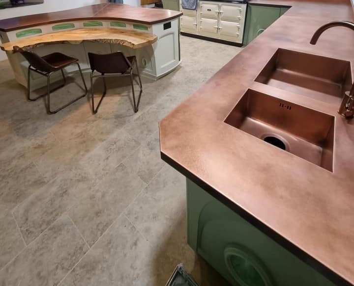 Copper kitchen worktop with two integrated sinks - Designs 05 Bespoke copper kitchen worktop with two seamlessly integrated copper sinks, featuring a warm aged patina finish. Paired with sage green cabinetry and a rustic breakfast bar, this custom copper design enhances both functionality and style in a luxury kitchen setting