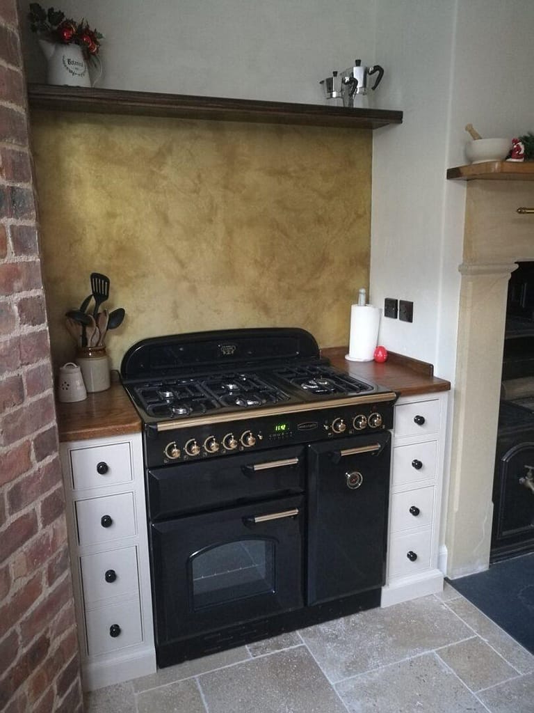 Lightly Aged Kitchen Splash Back - Designs 05 Example of brass splashbacks a lightly aged brass splashback with a rich patina, installed behind a traditional black range cooker, complemented by wooden worktops and exposed brick walls.