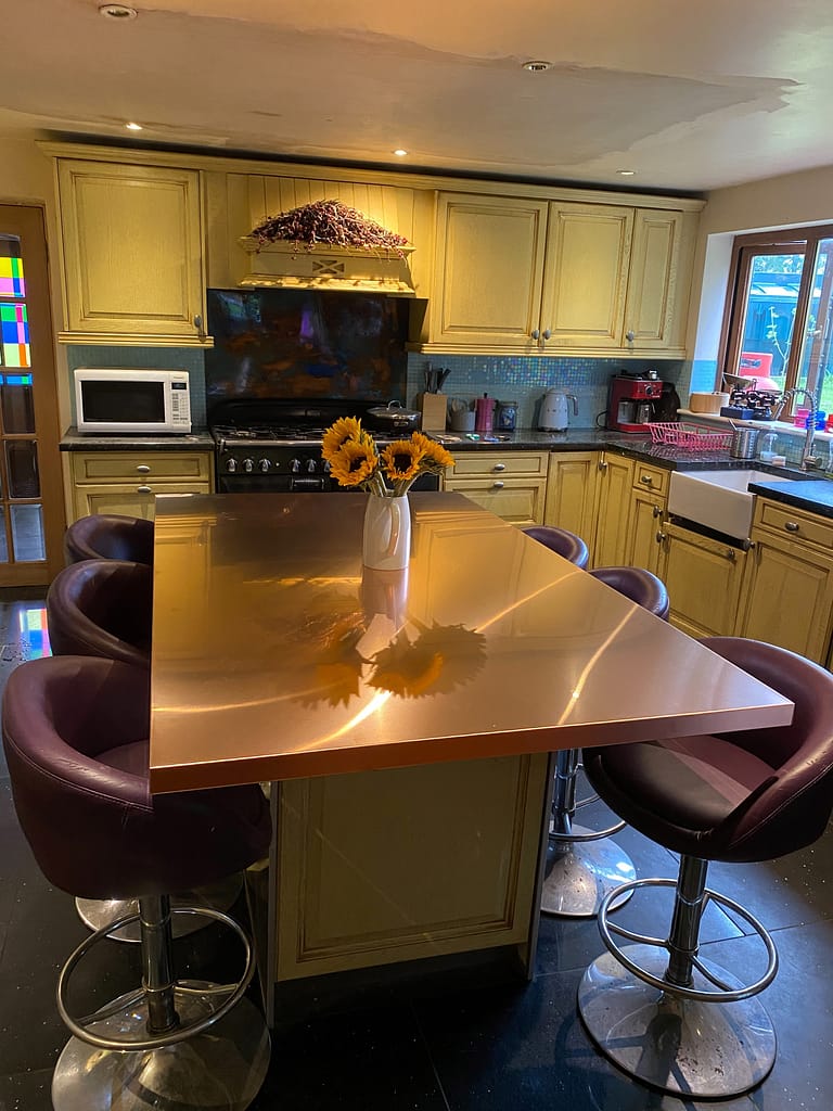 Copper Dining Table 4 seat - Designs 05 A six seater dining table with a copper top in a kitchen setting. A vase of flowers is on the top.
