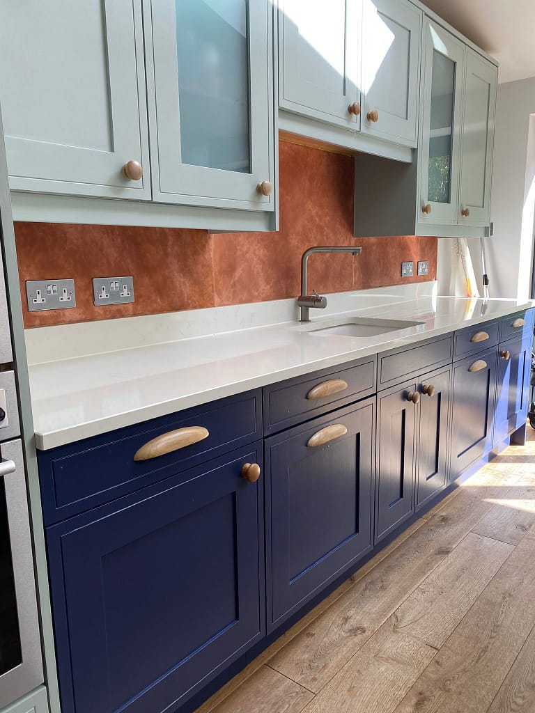 Copper splashback with four sockets - Designs 05 Bespoke aged copper kitchen splashback installed above a white quartz worktop, framed by navy and sage cabinetry, adding warmth and contrast to the modern kitchen design.
