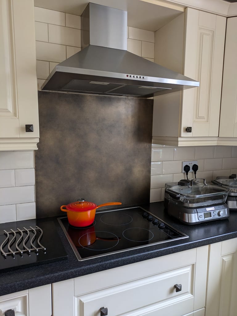 Antique Brass Splashback - Designs 05 An antique brass splashback installed behind a modern electric hob, paired with a stainless steel extractor hood and classic cream cabinetry.
