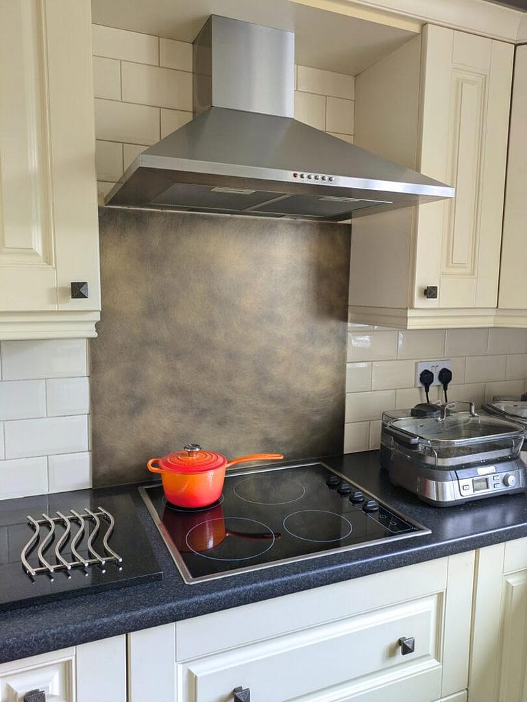 Burnished-Antique-Brass - Designs 05 Burnished version of the antique brass splashback installed behind a modern electric hob, paired with a stainless steel extractor hood and classic cream cabinetry.