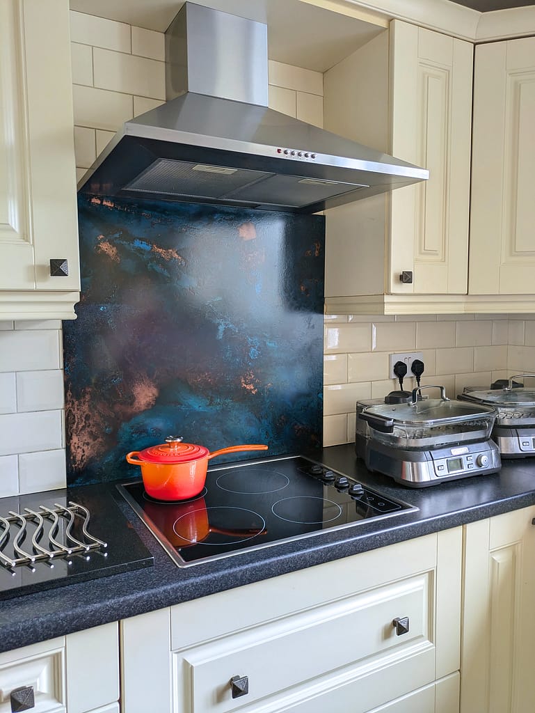 Blue Patina Copper Splashback – Aged & Textured Finish - Designs 05 Handcrafted blue patina copper splashback with rich aged texture behind hob in a cream kitchen with black countertops.