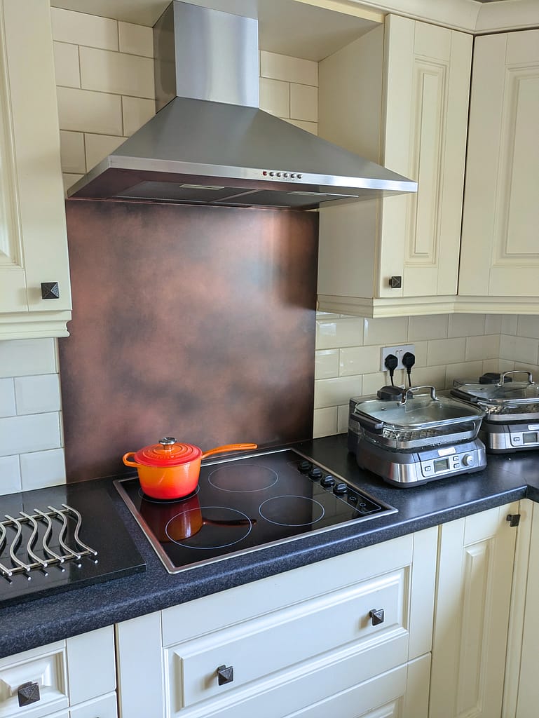 Dark Antique Copper Splashback with Cream Cabinets - Designs 05 Dark antique copper splashback installed behind a cooker with cream cabinets and black worktops.