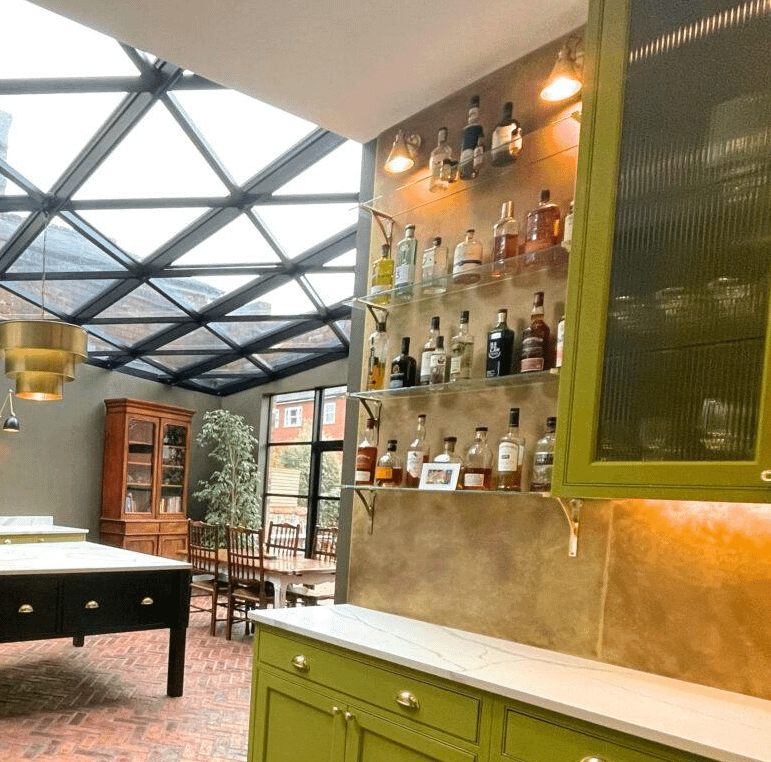 Lightly Aged Brass Splashback behind Drinks Shelves - Designs 05 Elegant lightly aged brass splashbacks featured in a home bar area, complementing glass shelves, brass accents, and green cabinetry for a sophisticated design